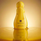 A bottle of Avon Her Wisdom Indulgent Glow Bath & Shower Oil, styled in a golden yellow color with dripping details, standing on a reflective surface against a warm gradient background.